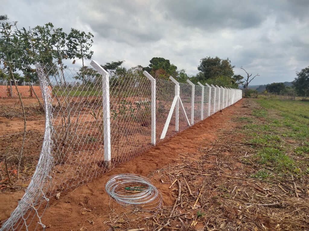 Trabalhador esticando o alambrado e fixando-o nos pilares alinhados, com um rolo de arame ao lado, representando um passo crucial na construção da cerca.