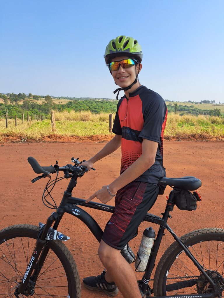 Joãozinho pedala na bicicleta de seu pai com entusiasmo, destacando o logo do Batman em seu quadro, em um gesto de puro orgulho e paixão pelo ciclismo.