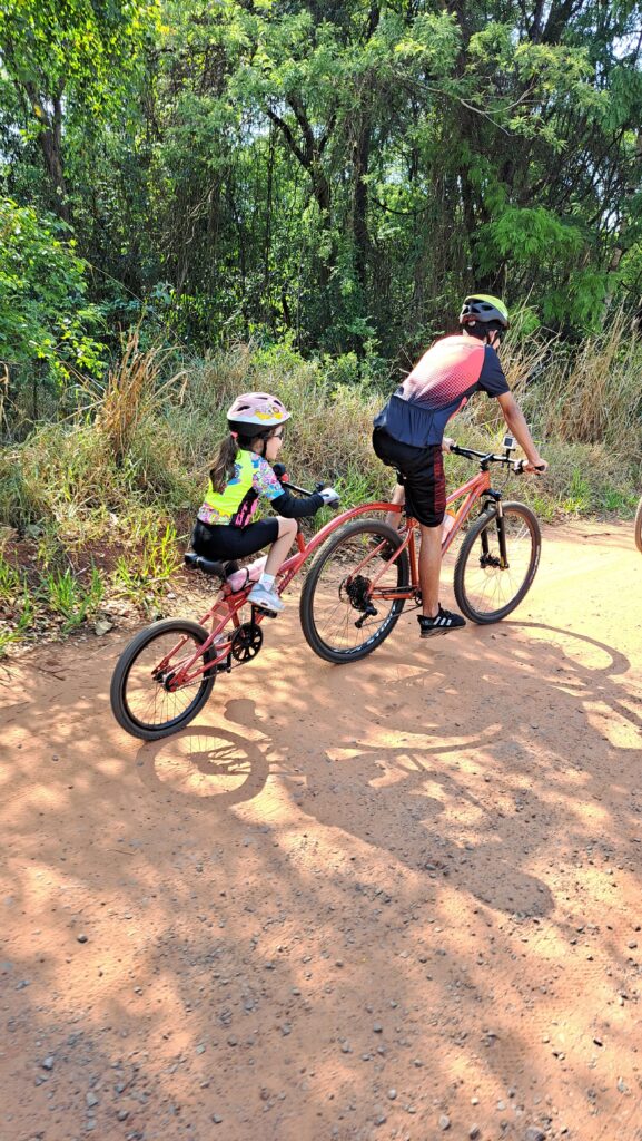 Joazinho pedala na bicicleta de seu tio, puxando o reboque onde Laurinha aproveita a jornada. Uma imagem que captura a alegria e a cumplicidade entre primos em uma emocionante aventura na Trilha do Bezerrão.