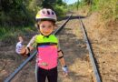 Laurinha, aventureira de 6 anos, sobre os trilhos do trem, fazendo um gesto de "joinha" enquanto aproveita sua jornada de bicicleta.