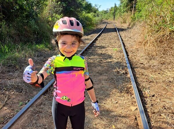 Laurinha, aventureira de 6 anos, sobre os trilhos do trem, fazendo um gesto de "joinha" enquanto aproveita sua jornada de bicicleta.